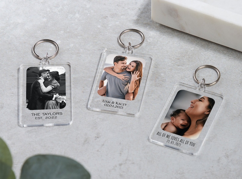 May include: Three clear acrylic keychains with photos and text. The first keychain has a black and white photo of a couple and the text "THE TAYLORS EST. 2022". The second keychain has a color photo of a couple and the text "LIAM & KACEY 02.04.2023". The third keychain has a color photo of a mother and child and the text "ALL OF ME LOVES ALL OF YOU 23.03.2023".