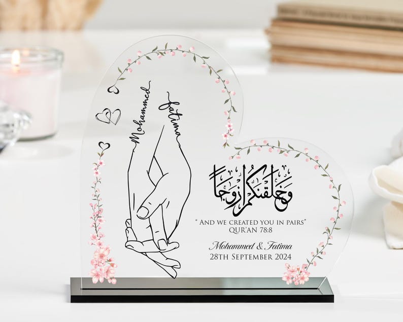 May include: A clear acrylic heart-shaped plaque with pink floral designs and the names "Mohammed" and "Fatima" written in calligraphy. The plaque also includes the text "And we created you in pairs" and a Quran verse reference. The date "28th September 2024" is also included.