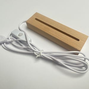 Puede incluir: Una base de luz LED rectangular de madera con una ranura para exhibir objetos. Tiene un cable USB blanco con un interruptor de encendido/apagado en línea. La base es de madera de color claro.