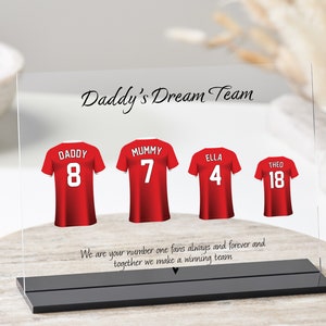 K&ouml;nnte beinhalten: Eine klare Acrylplatte mit schwarzem Sockel, die ein rot-wei&szlig;es Fu&szlig;balltrikot-Design mit den Worten "Daddy's Dream Team" und den Namen "Daddy", "Mummy", "Ella" und "Theo" mit entsprechenden Trikotnummern zeigt. Der Text unter den Trikots lautet "We are your number one fans always and forever and together we make a winning team."