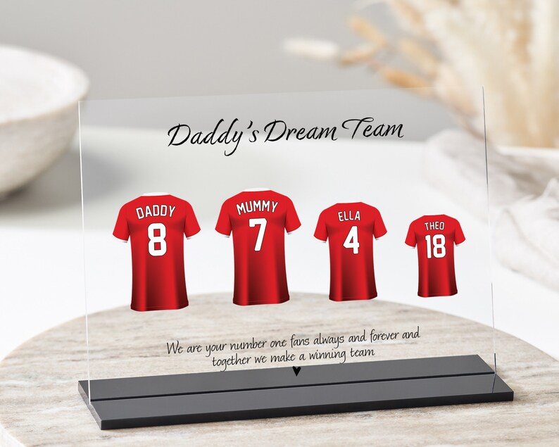 K&ouml;nnte beinhalten: Eine klare Acrylplatte mit schwarzem Sockel, die ein rot-wei&szlig;es Fu&szlig;balltrikot-Design mit den Worten "Daddy's Dream Team" und den Namen "Daddy", "Mummy", "Ella" und "Theo" mit entsprechenden Trikotnummern zeigt. Der Text unter den Trikots lautet "We are your number one fans always and forever and together we make a winning team."
