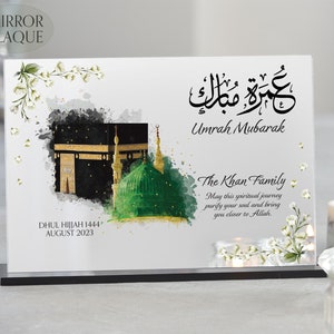 May include: A white mirror plaque with a watercolor image of the Kaaba and the Green Dome in Mecca. The plaque has the Arabic text "Umrah Mubarak" and the text "The Khan Family" with a message wishing them a blessed Umrah. The plaque also includes the date "Dhul Hijjah 1444, August 2023".