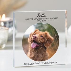 Può includere: Un blocco commemorativo in acrilico trasparente con una foto di un cane marrone. Il testo "Bella" è in alto, seguito da "You left a paw print on our hearts 2012-2023". La parte inferiore del blocco recita "FOREVER loved, NEVER forgotten".