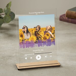 Może przedstawiać: Przezroczysty akrylowy wyświetlacz fotograficzny z drewnianą podstawą. Zdjęcie przedstawia trzy osoby na polu słoneczników, z tekstem "Personal Message Here" i fioletowym graficznym kształtem fali dźwiękowej. Wyświetlany jest również tytuł piosenki "Count On Me" Bruno Marsa.