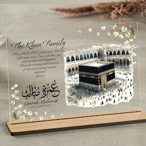 Puede incluir: Una placa de acrílico transparente con un diseño floral blanco y una ilustración en blanco y negro de la Kaaba. La placa tiene el texto "The Khan Family" y "Umrah Mubarak December 2024" en letras negras.