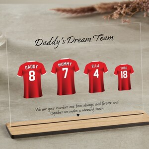 K&ouml;nnte beinhalten: Eine durchsichtige Acrylplatte mit Holzsockel, die ein rotes Fu&szlig;balltrikot-Design mit dem Text "Daddy's Dream Team" und den Namen "Daddy", "Mummy", "Ella" und "Theo" mit entsprechenden Nummern zeigt. Der Text unter den Trikots lautet "We are your number one fans always and forever and together we make a winning team."