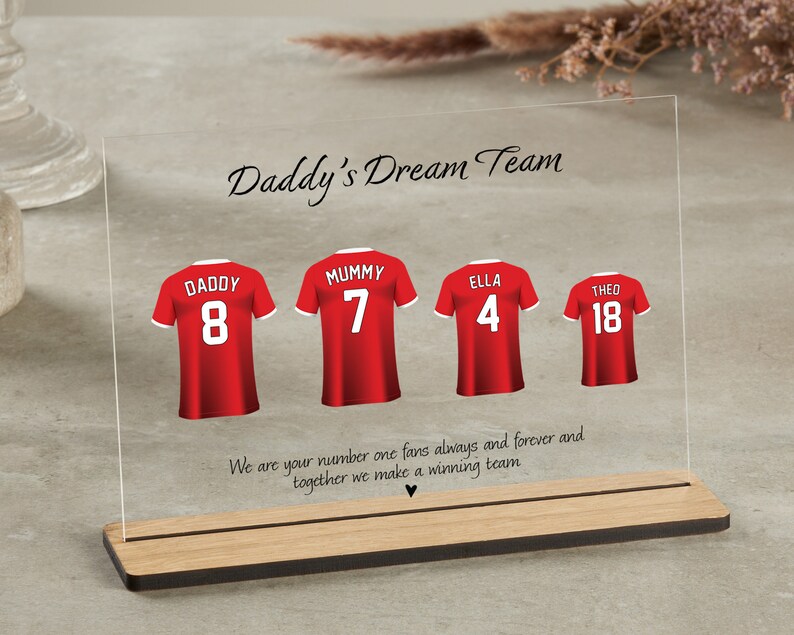 K&ouml;nnte beinhalten: Eine durchsichtige Acrylplatte mit Holzsockel, die ein rotes Fu&szlig;balltrikot-Design mit dem Text "Daddy's Dream Team" und den Namen "Daddy", "Mummy", "Ella" und "Theo" mit entsprechenden Nummern zeigt. Der Text unter den Trikots lautet "We are your number one fans always and forever and together we make a winning team."