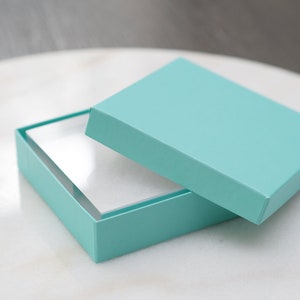 May include: A light blue gift box with a white interior. The lid is open and the box is sitting on a white surface.
