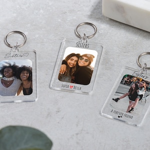Op de afbeelding: Drie transparante acryl sleutelhangers met zilveren sleutelringen. Elke sleutelhanger heeft een rechthoekige foto van lachende mensen. Het woord "Sisters" staat bovenaan elke sleutelhanger. Eén sleutelhanger zegt "A Forever Friend".