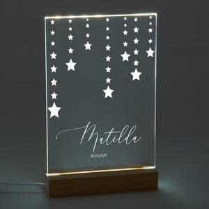 Pode incluir: Uma luz noturna de acrílico branco com uma base de madeira. A luz apresenta um design de estrelas brancas penduradas em cordas. O nome "Matilda" e a data "26.04.2024" também estão incluídos no design.