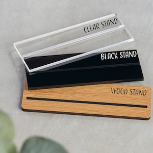 May include: Three different display stands for a sign or plaque. The stands are made of clear acrylic, black acrylic, and wood. The stands are labeled "CLEAR STAND", "BLACK STAND", and "WOOD STAND".