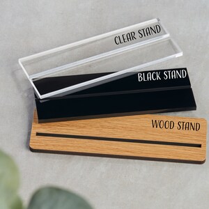 K&ouml;nnte beinhalten: Drei Pr&auml;sentationsst&auml;nder f&uuml;r ein Schild oder eine Plakette. Die St&auml;nder sind aus klarem Acryl, schwarzem Acryl und Holz. Die St&auml;nder sind mit "CLEAR STAND", "BLACK STAND" und "WOOD STAND" beschriftet.