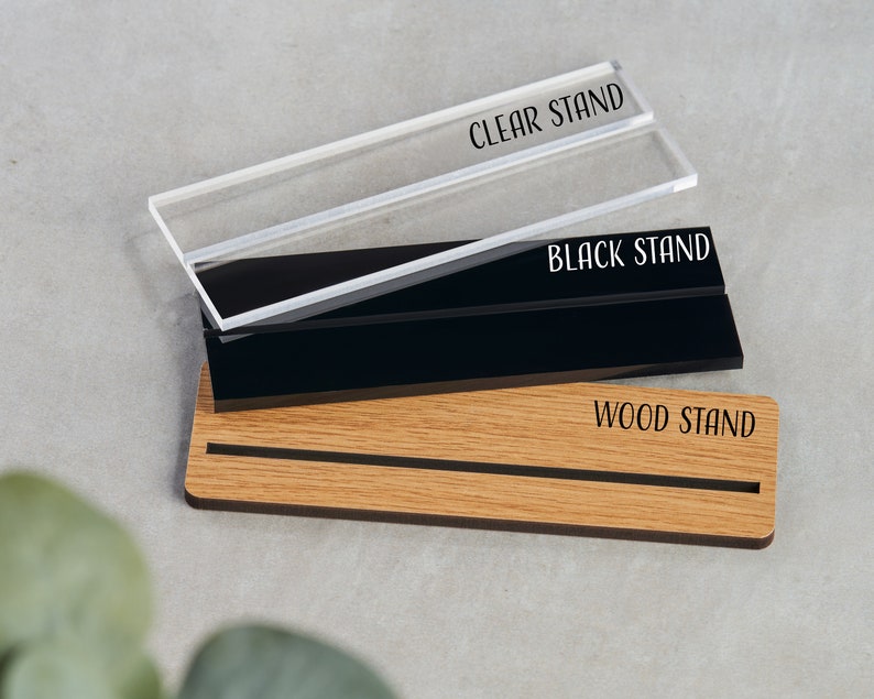 K&ouml;nnte beinhalten: Drei Pr&auml;sentationsst&auml;nder f&uuml;r ein Schild oder eine Plakette. Die St&auml;nder sind aus klarem Acryl, schwarzem Acryl und Holz. Die St&auml;nder sind mit "CLEAR STAND", "BLACK STAND" und "WOOD STAND" beschriftet.