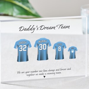 K&ouml;nnte beinhalten: Ein klarer Acrylblock mit dem Text "Daddy's Dream Team" darauf gedruckt. Es gibt vier blaue und wei&szlig;e Trikots mit darauf gedruckten Nummern und Namen. Die Trikots sagen "Daddy 32", "Mummy 30", "Leah 8" und "Mild". Der Text unter den Trikots lautet "We are your number one fans always and forever and together we make a winning team."