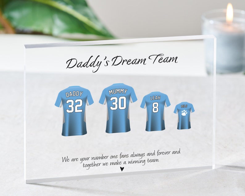 K&ouml;nnte beinhalten: Ein klarer Acrylblock mit dem Text "Daddy's Dream Team" darauf gedruckt. Es gibt vier blaue und wei&szlig;e Trikots mit darauf gedruckten Nummern und Namen. Die Trikots sagen "Daddy 32", "Mummy 30", "Leah 8" und "Mild". Der Text unter den Trikots lautet "We are your number one fans always and forever and together we make a winning team."
