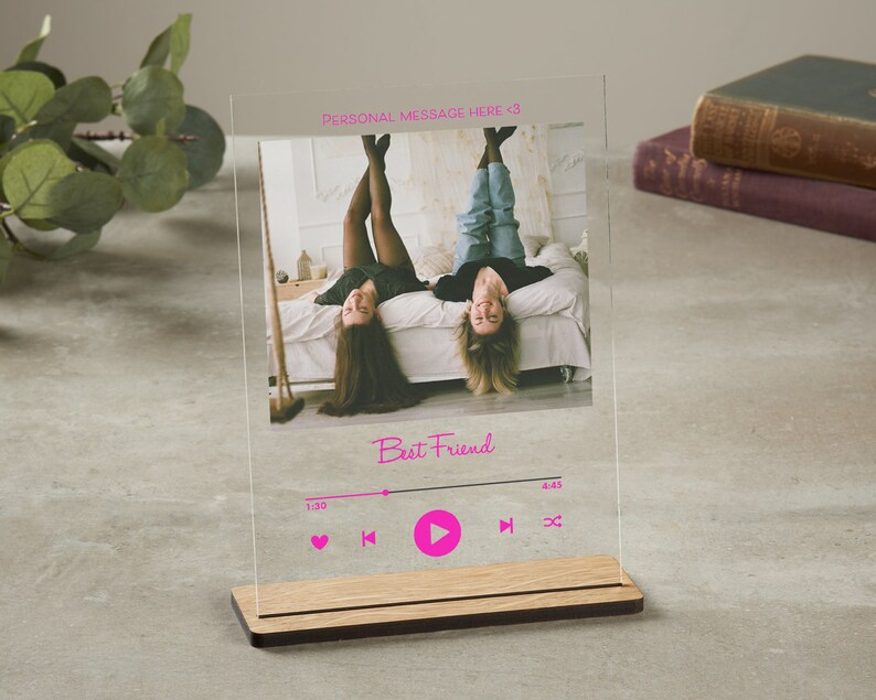 May include: A clear acrylic photo frame with a wood base. The frame features a photo of two friends lying on a bed with their legs in the air. The frame has a music player design with the text "Best Friend" and play, pause, and skip buttons.
