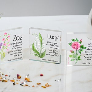 May include: Three clear acrylic blocks with watercolour floral designs and personalised messages. The first block features a pink and purple flower with the name "Zoe" and the message "Forget the past; look forward to the future, for the best things are get to come. From Grace. Blessed | Grateful | Reliable". The second block features a white lily with the name "Lucy" and the message "Wishing you a day filled with love and a year of joy. Lots of love Mum and Dad. Humble | Sweet | Beautiful". The third block features a pink rose with the name "Ella" and the message "Life is better with someone like you in it. Wishing you joy and sunshine in the coming year. From Melissa. Loving | Fair | Passionate".