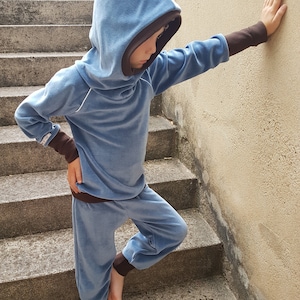 May include: A child wearing a blue hooded sweatshirt and matching blue sweatpants with brown trim. The child is standing on a set of concrete stairs.