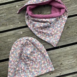 May include: A set of two floral patterned fabric items: a beanie and a bandana. The beanie is a light pink color with a floral pattern in shades of blue, green, and purple. The bandana is a dark purple color with a floral pattern in shades of blue, green, and purple.