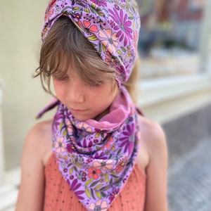 Könnte beinhalten: Ein floraler Haarband- und Halstuch-Set. Das Set zeigt einen lavendelfarbenen Hintergrund mit rosa, orangefarbenen und lila Blumen und grünen Blättern. Das Haarband ist verdreht und das Halstuch ist dreieckig.