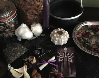 May include: A collection of items for spiritual practices, including a black cauldron, a purple candle, garlic cloves, almonds, a sea urchin shell, and a tarot card featuring a three-faced figure. Jars of herbs and dried flowers are also present.