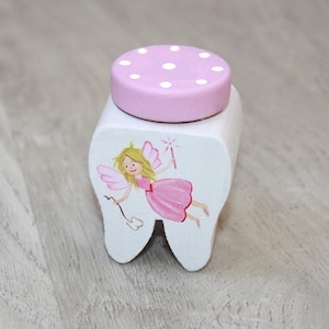 Könnte beinhalten: Eine weiße, zahnförmige Box mit einem rosa Deckel mit weißen Punkten. Die Box ist mit einem gemalten Bild einer Fee mit rosa Flügeln und einem rosa Kleid verziert.