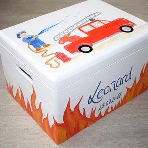 May include: White wooden toy chest with a fire truck, firefighter, and flames painted on the side. The chest has a handle and the name "Leonard" and the date "27.07.2047" are written on the side.