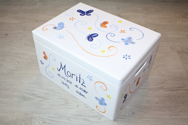 May include: White wooden storage box with a blue, orange, and yellow butterfly design. The box is decorated with the name "Moritz" and the date "15.04.2020 23:45 46.15cm 3950g".