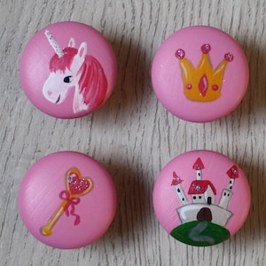 Könnte beinhalten: Vier runde, rosa Schubladenknöpfe mit handgemalten Motiven. Ein Knopf zeigt ein weißes Einhorn, ein weiterer eine goldene Krone, der dritte einen goldenen herzförmigen Zauberstab mit Glitzer und der vierte ein weißes Schloss mit roten Dächern.