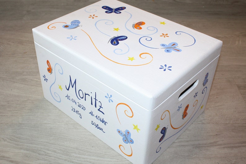 May include: White wooden keepsake box with a blue and orange butterfly design. The box is personalized with the name "Moritz" and the date "15.04.2020".