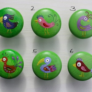 Könnte beinhalten: Sechs grüne Holz-Knöpfe mit bemalten Vogelmotiven. Jeder Knopf zeigt einen anderen Vogel mit einzigartigen Farben und Mustern. Die Knöpfe sind von 1 bis 6 nummeriert.