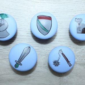 Könnte beinhalten: Fünf runde blaue Knöpfe mit verschiedenen mittelalterlichen Designs, die darauf gemalt sind. Die Designs umfassen einen Ritterhelm, einen Schild, ein Schloss, ein Schwert und eine Dornenkeule.