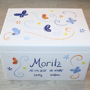 May include: White wooden keepsake box with a blue, orange, and yellow butterfly design. The box is decorated with swirls and stars. The text "Moritz 15.04.2020 16:15Uhr 2945g 50,5cm" is written on the front of the box.
