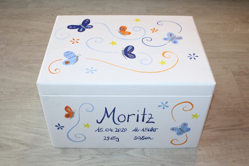 May include: White wooden keepsake box with a blue, orange, and yellow butterfly design. The box is decorated with swirls and stars. The text "Moritz 15.04.2020 16:15Uhr 2945g 50,5cm" is written on the front of the box.