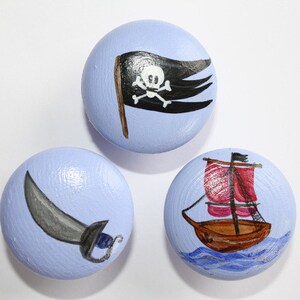 Könnte beinhalten: Drei Holz-Knöpfe mit Piraten-Motiv bemalt. Ein Knopf zeigt eine schwarze Piratenflagge mit einem weißen Totenkopf und Knochen. Ein weiterer Knopf hat ein graues Piratenschwert. Der dritte Knopf zeigt ein rot-braunes Segelboot mit einem roten Segel.