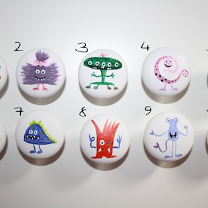 Könnte beinhalten: Zehn runde weiße Keramikknöpfe mit verschiedenen bunten Monsterdesigns, die darauf gemalt sind. Die Knöpfe sind von 1 bis 10 nummeriert.