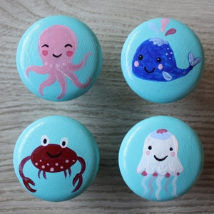 Könnte beinhalten: Vier runde Holzknöpfe, die mit einem hellblauen Hintergrund und verschiedenen Meeresbewohnern bemalt sind. Die Knöpfe zeigen einen Oktopus, einen Wal, einen Krebs und eine Qualle.