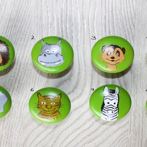 Könnte beinhalten: Acht grüne Holzgriffe mit verschiedenen Tiergesichtern, die darauf gemalt sind. Die Tiere sind ein Löwe, ein Flusspferd, ein Erdmännchen, eine Giraffe, ein Elefant, ein Tiger, ein Zebra und ein Affe.