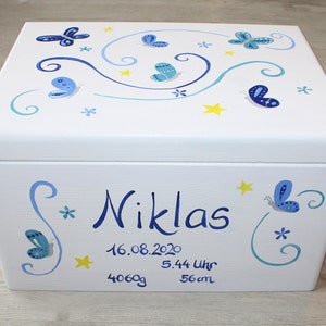 May include: White wooden keepsake box with blue butterflies and yellow stars painted on the lid. The box is personalized with the name "Niklas", the date "16.08.2020", the time "5.44 Uhr", the weight "4060g", and the length "56cm".