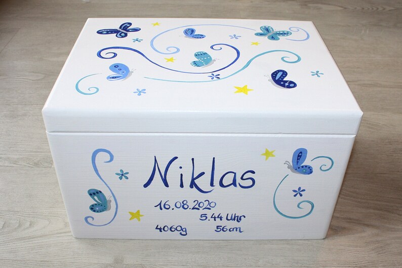 May include: White wooden keepsake box with blue butterflies and yellow stars painted on the lid. The box is personalized with the name "Niklas", the date "16.08.2020", the time "5.44 Uhr", the weight "4060g", and the length "56cm".