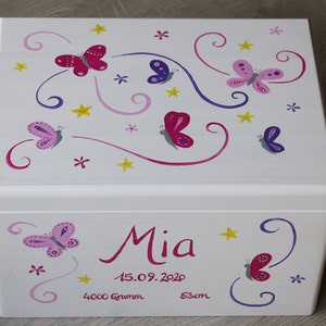 May include: White wooden keepsake box with a butterfly theme. The lid is decorated with pink, purple, and yellow butterflies and swirls. The box is personalized with the name "Mia", the date "15.09.2020", and the measurements "4000 Gramm" and "53cm".