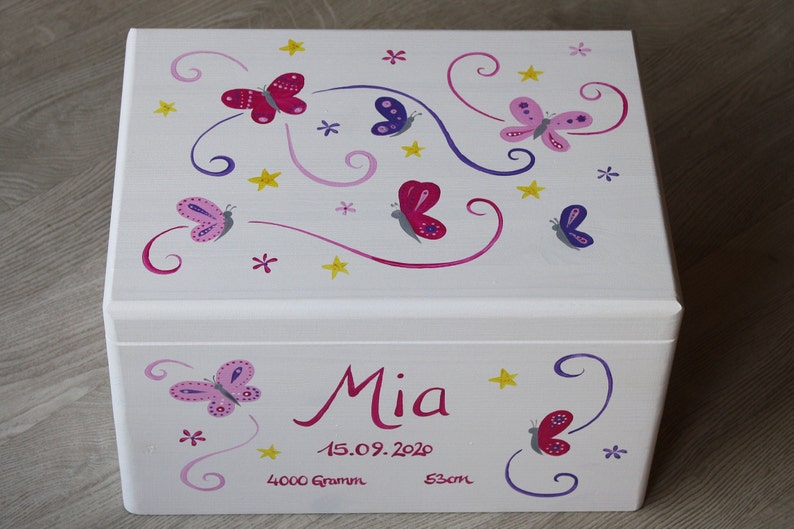 May include: White wooden keepsake box with a butterfly theme. The lid is decorated with pink, purple, and yellow butterflies and swirls. The box is personalized with the name "Mia", the date "15.09.2020", and the measurements "4000 Gramm" and "53cm".