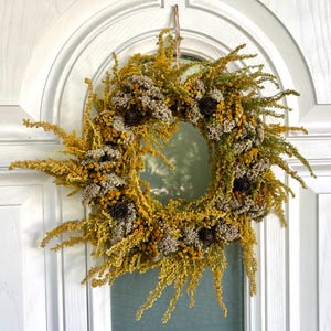 May include: A circular wreath made of dried yellow and green foliage, accented with small brown pinecones and beige flowers. The wreath is hanging on a white door with a glass window.