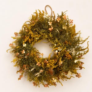 May include: A circular wreath composed of dried foliage in shades of mustard yellow, olive green, and muted brown. The arrangement features textured sprigs, small clusters of golden yellow flowers, and delicate pale petals, with a jute loop at the top.