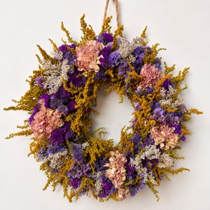 Könnte beinhalten: Ein kreisförmiger Kranz aus getrockneten Blumen und Pflanzen. Das Arrangement besteht aus Büscheln von blassrosa Hortensienblüten, leuchtend violettem Statice, goldgelbem Solidago und Zweigen von silbrig-weißen Trockenpflanzen.