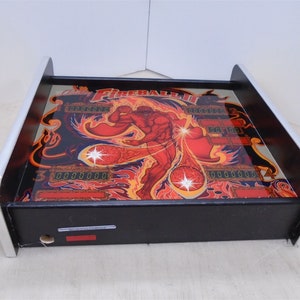May include: A black and silver pinball machine with a red, orange, and yellow graphic of a man with a fireball. The machine has a digital display that reads "0000000".