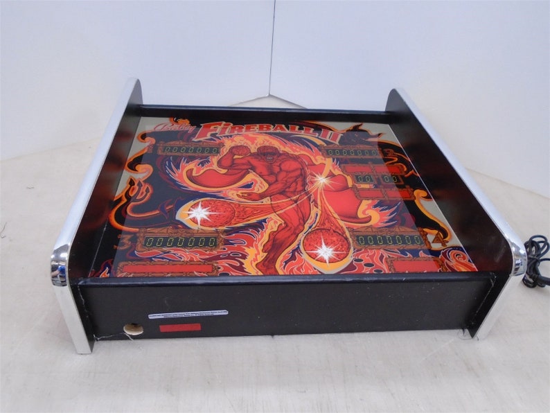 May include: A black and silver pinball machine with a red, orange, and yellow graphic of a man with a fireball. The machine has a digital display that reads "0000000".