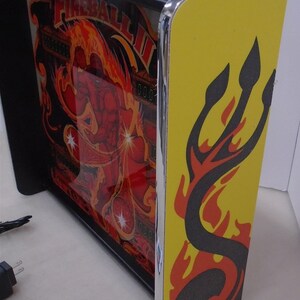 May include: A black framed light-up pinball machine with a red and yellow image of a man in flames. The machine has a yellow side panel with a black and red flame design.