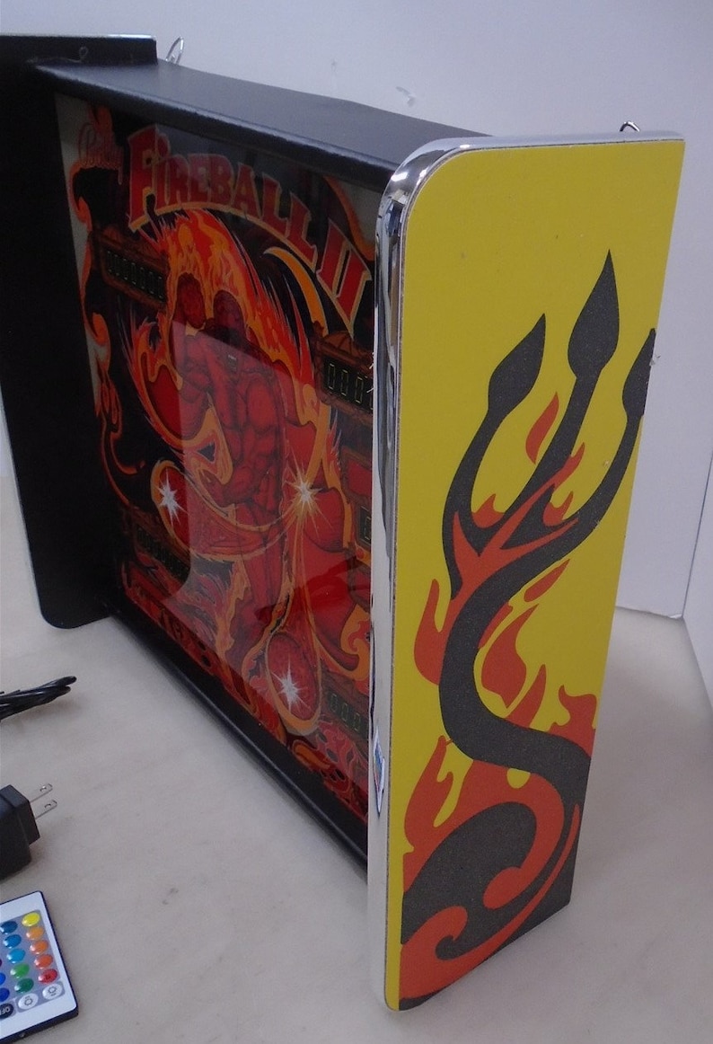 May include: A black framed light-up pinball machine with a red and yellow image of a man in flames. The machine has a yellow side panel with a black and red flame design.