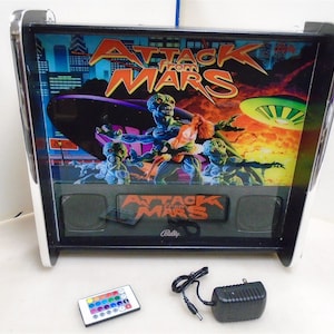Puede incluir: Una máquina de pinball en miniatura con un gabinete blanco y negro y un cristal trasero colorido con el texto "Attack from Mars" y una representación de extraterrestres. La máquina incluye un control remoto y un adaptador de corriente.